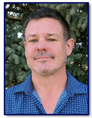 A man with a blue shirt is standing in front of some trees